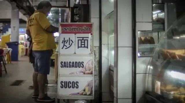 Imagem de lanchonete dentro do Mercado Central de Camaçari com foco em uma placa com os dizeres "Promoção, salgados, sucos, junto de ideogramas em mandarim".