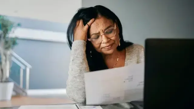 Uma mulher estressada está sentada à mesa em casa, lendo um documento ao lado do laptop. Ela parece preocupada e tensa, com a cabeça apoiada nas mãos enquanto administra suas contas pessoais.
